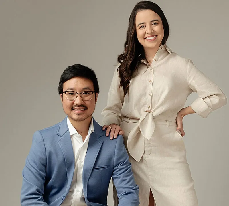 Dr. Guilherme Massami Horie sentado e Dra. Larissa Negrão Ferreira em pé ao lado dele, os dois sorrindo, em retrato de estúdio sobre fundo taupe