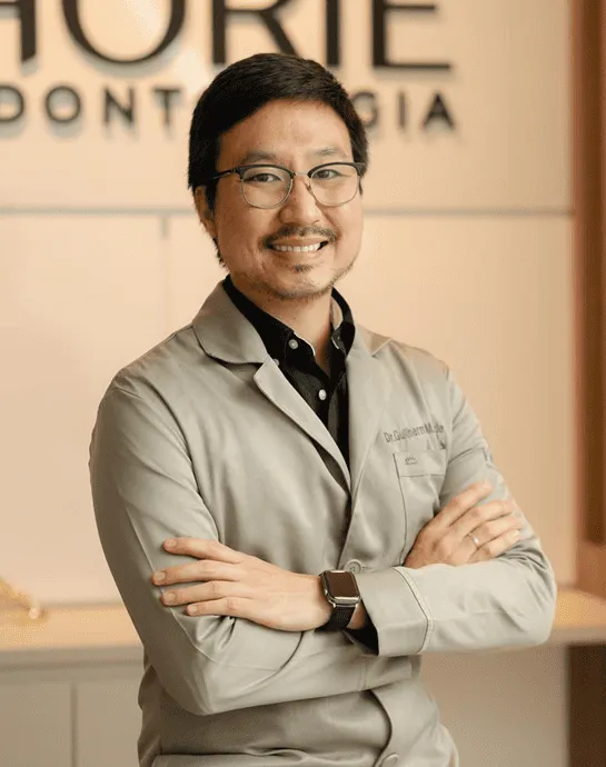 Dr. Guilherme Massami Horie de jaleco, braços cruzados, sorrindo, com o logotipo da clínica na parede ao fundo