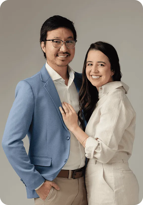 Dr. Guilherme Massami Horie e Dra. Larissa Negrão Ferreira lado a lado, sorrindo, em retrato de estúdio sobre fundo taupe