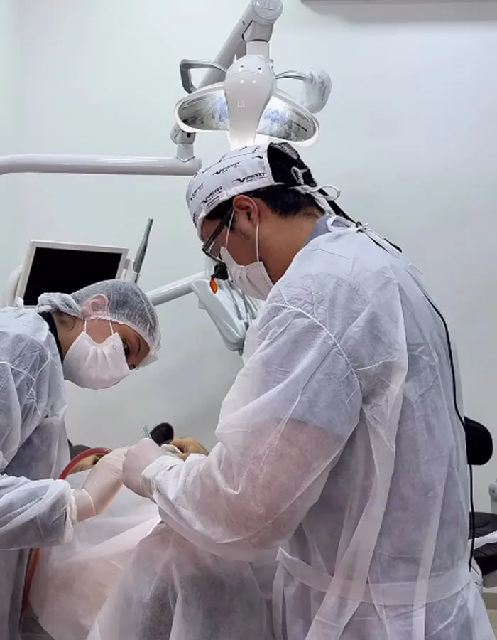 Dois cirurgiões-dentistas de avental, touca, máscara e luvas atendendo um paciente sob o refletor do consultório